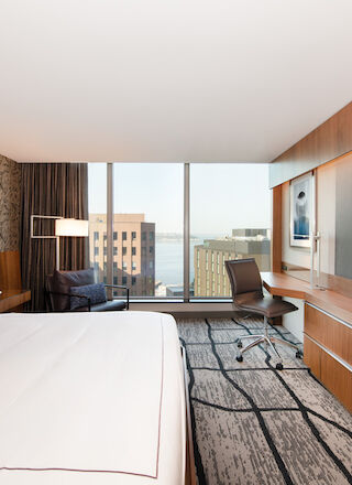 A modern hotel room has a bed, a large TV, a desk with a chair, a corner chair, and a window with a city view, including water and buildings.