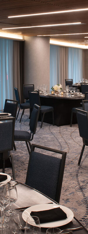 The image shows an elegant banquet room with round tables set for an event, featuring centerpieces, glassware, and folded napkins on a patterned carpet.