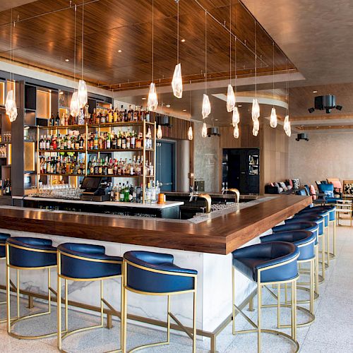 The image shows a modern bar with wooden elements, blue barstools, hanging lights, and a well-stocked liquor shelf, located in a stylish, spacious lounge area.