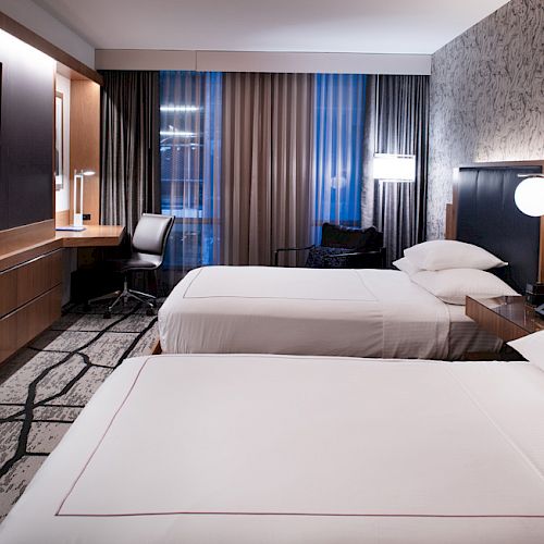 A modern hotel room with two neatly made beds, a wall-mounted TV, a desk with a chair, and large windows with curtains.
