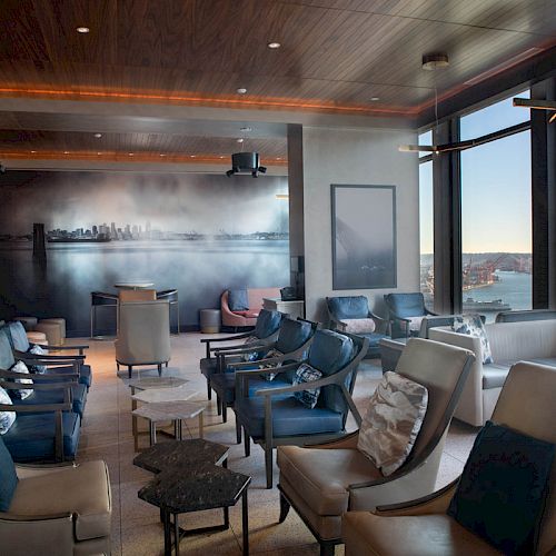 A modern lounge with plush seating, large windows offering a scenic waterfront view, and wall art depicting a city skyline is shown.