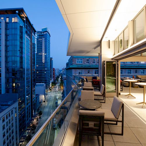 View from an upscale balcony with modern furniture overlooking a city street lined with high-rise buildings at dusk.