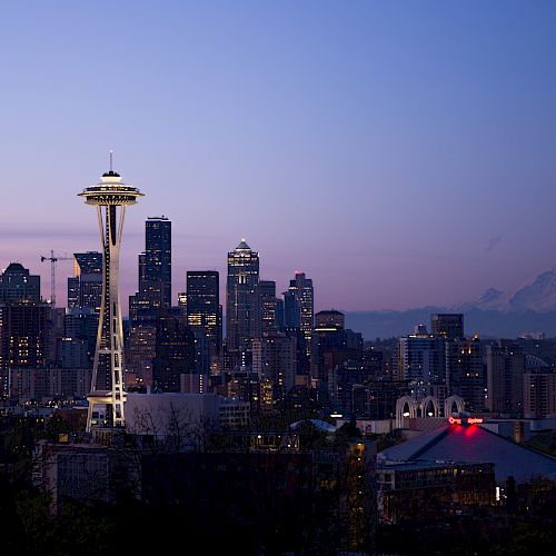 This image shows the skyline of Seattle at dusk, featuring the Space Needle and Mount Rainier in the background, with city lights starting to illuminate.