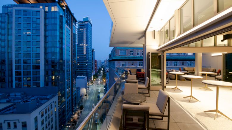 The image shows a modern urban balcony at night, with outdoor seating and a view of high-rise buildings and a street below.