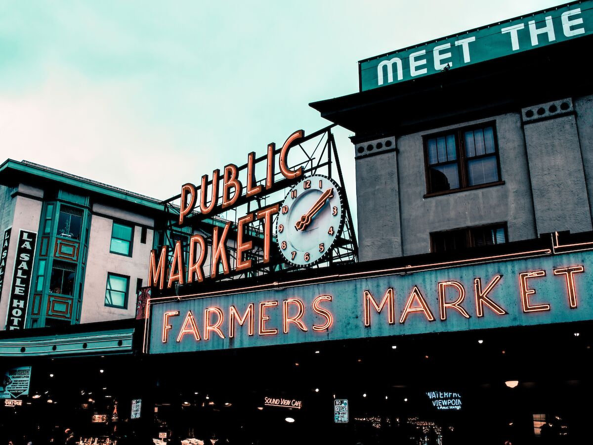 A neon sign for "Public Market" and "Farmers Market," featuring a clock, set against an urban building backdrop likely indicating a bustling market area.