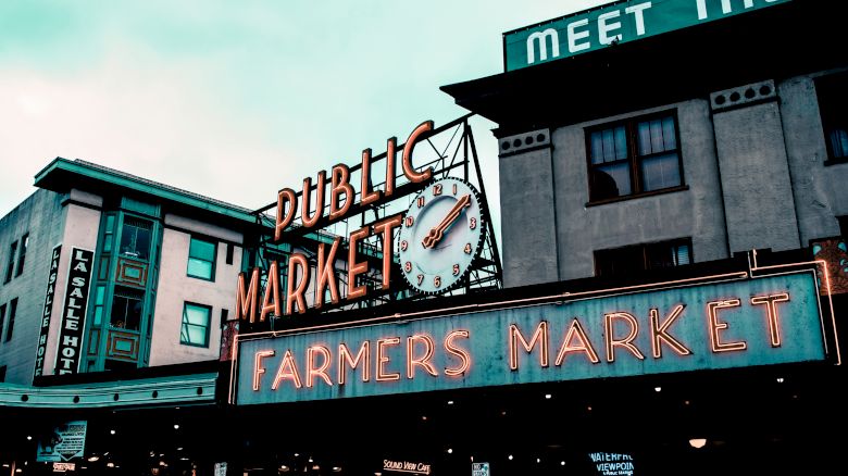A neon sign for "Public Market" and "Farmers Market," featuring a clock, set against an urban building backdrop likely indicating a bustling market area.