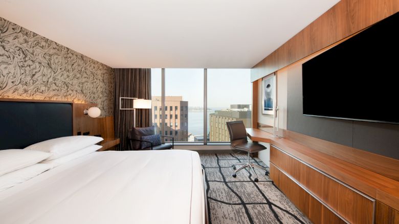 A modern hotel room with a large bed, a desk, a chair, a flat-screen TV, and a window offering a city view.