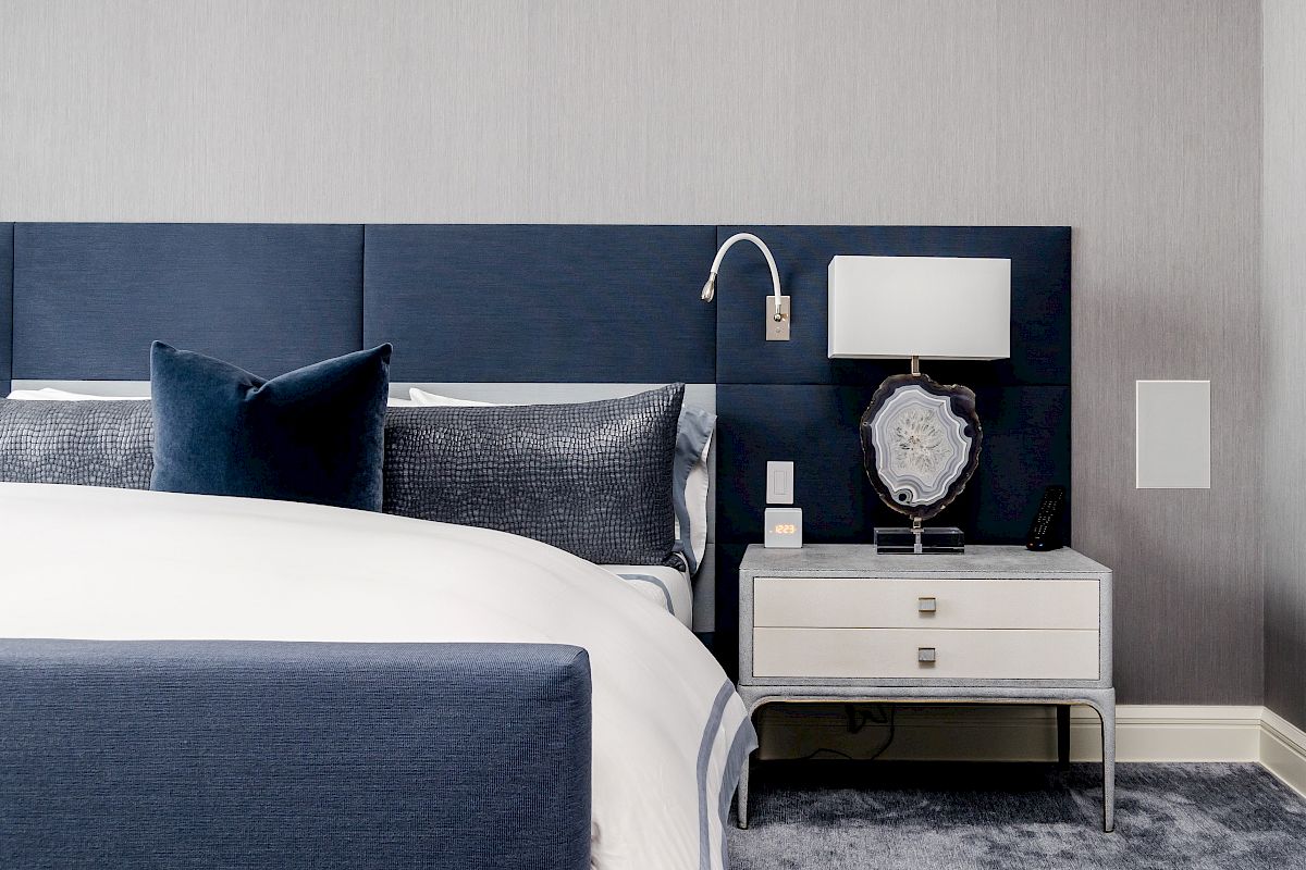 A modern bedroom with a navy blue headboard, white bedding, a nightstand with a lamp, and a decorative item on it, all on a plush gray carpet.