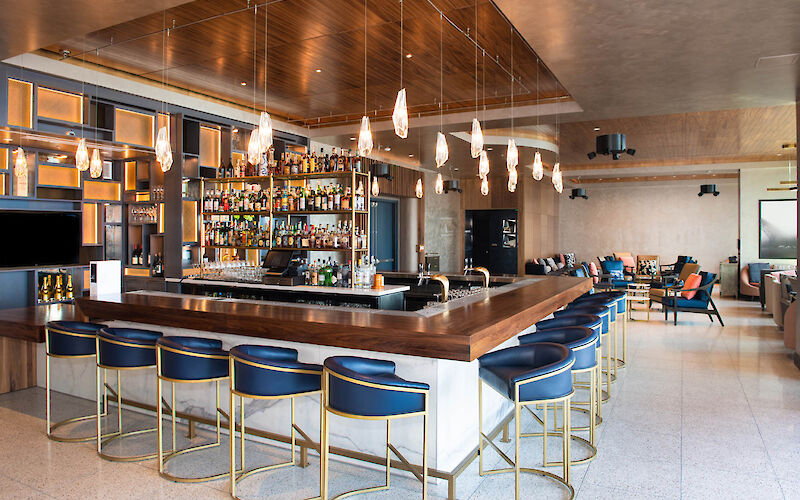 The image shows a modern bar with blue barstools, a well-stocked liquor shelf, pendant lights, and a lounge area with couches and chairs in the background.