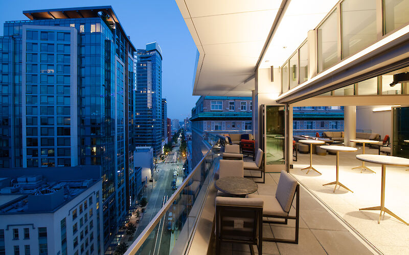 The image shows a modern balcony with tables and chairs overlooking a cityscape with tall buildings at dusk.