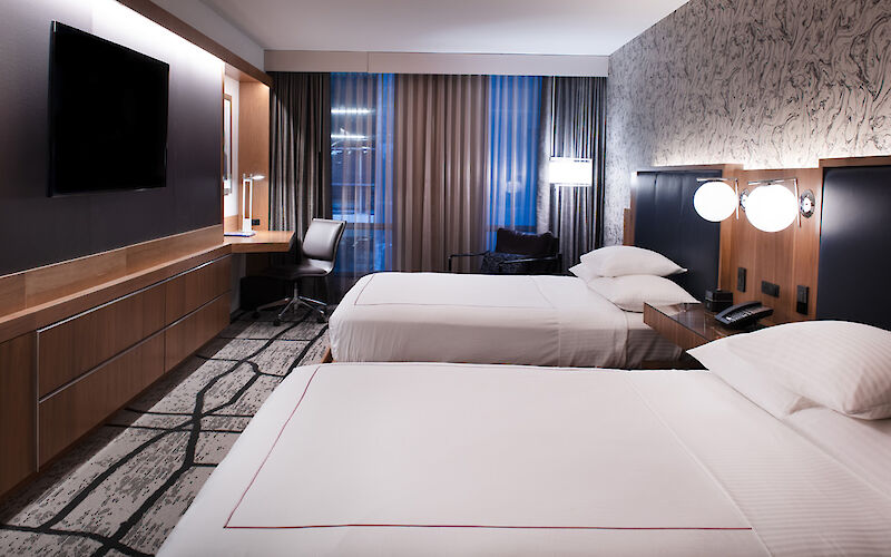 A modern hotel room with two neatly made beds, a flat-screen TV, a desk with a chair, and elegant decor, including a patterned wall.