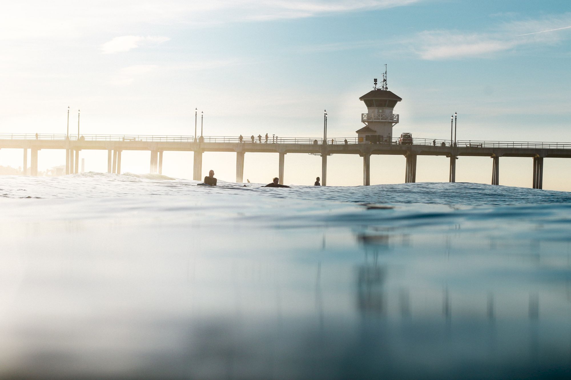 A pier stretches across calm waters, with a few people and surfers visible in the distance under a blue sky.